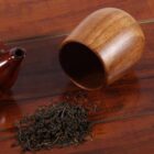 Wooden cup tipped over on a wooden table with a small pile of loose leaf tea spilling out, next to a brown teapot lid.