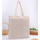 A plain beige cloth tote bag with handles is set on a white surface. In the background, there are wicker baskets and some green plants, creating a simple and natural aesthetic.