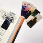 A book is open to page 8 with a RiverClan themed magnetic bookmark attached. Nearby, four more bookmarks feature different nature and cat silhouettes with scenic backgrounds in various colors.
