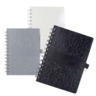 Three spiral-bound notebooks with textured covers in black, grey, and white are arranged standing upright. Each notebook features a black elastic band for closure.