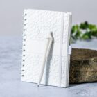A white textured notebook with a pen attached, placed on a stone surface. The background includes a blurred plant, giving a minimalist and natural feel.