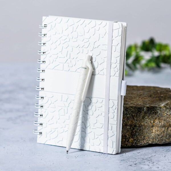 A white textured notebook with a pen attached, placed on a stone surface. The background includes a blurred plant, giving a minimalist and natural feel.