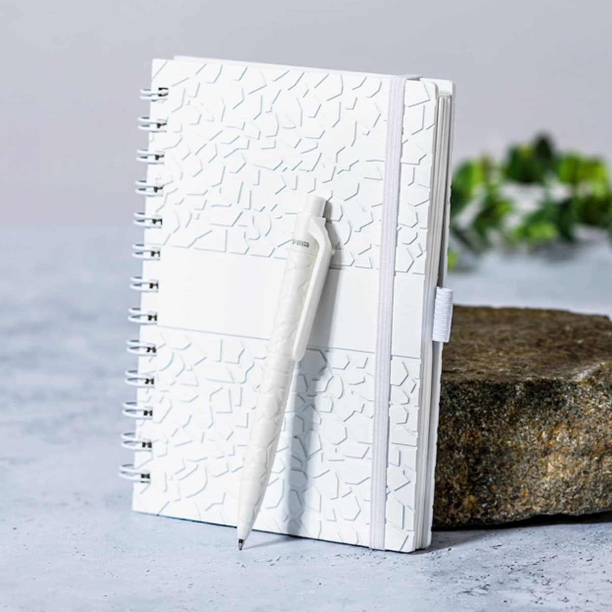 A white textured notebook with a spiral binding is placed on a rock surface. A matching white pen is clipped to the notebook. Some greenery is blurred in the background.
