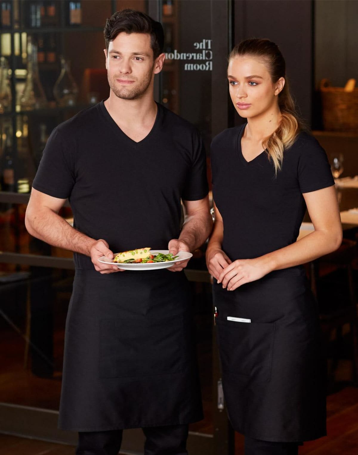 Two servers in black uniforms stand side by side. The person on the left holds a plate of food. They are in a restaurant setting with wine bottles visible in the background.