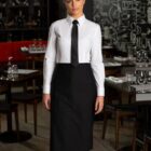 A person wearing a white shirt, black tie, and black apron stands in a restaurant with neatly arranged tables and a chalkboard wall in the background.