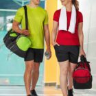 Two people in athletic gear walk indoors, smiling at each other. Both wear black caps, shorts, and sneakers. The person on the left wears a lime green shirt and carries a matching bag. The person on the right wears a red shirt and carries a matching bag.