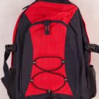 A red and black backpack with a handle and adjustable shoulder straps. It features multiple zippered compartments and two mesh side pockets. The front has a crisscross bungee cord detail.