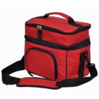 A red insulated lunch bag with a black zipper, black handle, and adjustable shoulder strap. It features a front pocket and a main compartment. The bag stands upright against a white background.