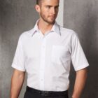 A man with short brown hair is wearing a white short-sleeve dress shirt and black trousers. He has a black belt and a watch, standing against a gray concrete wall, looking slightly to the side with a neutral expression.