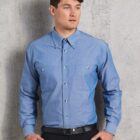 A person wearing a blue button-up shirt and black pants stands against a gray concrete wall. They have short, dark hair and are looking to their left. Their left hand is in their pocket, and they wear a black belt.