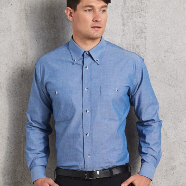A person wearing a blue button-up shirt with flap pockets and black trousers stands against a gray textured background. They are looking off to the side with a neutral expression, one hand in a pocket.