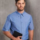 A man in a short-sleeve blue button-up shirt stands against a gray background, holding a black folder. He has short brown hair and a beard, and is looking slightly to the side.