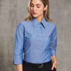 A woman with shoulder-length hair is wearing a blue button-up shirt with three-quarter sleeves and black pants. She has her right hand in her pocket and is standing against a textured gray background, looking downwards.