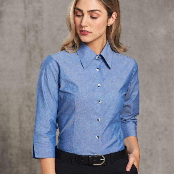 A woman with shoulder-length hair is wearing a blue button-up shirt and black pants with a belt. She stands against a gray textured background, looking down with her hand in her pocket.