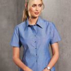 Woman with blond hair in a ponytail, wearing a blue short-sleeve button-up shirt and a watch, standing against a grey background.