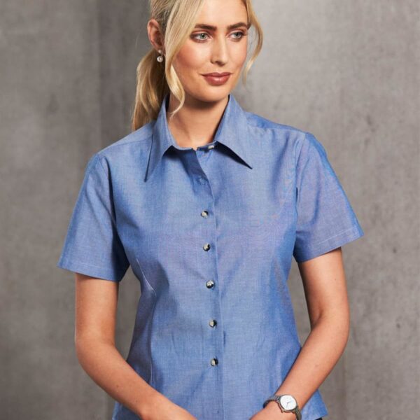 A woman with long blonde hair is wearing a blue short-sleeved button-up shirt and dark pants, standing against a plain gray background. She has a watch on her left wrist and pearl earrings, looking to her left with a neutral expression.