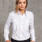 A person wearing a white button-up shirt with epaulets and black pants stands against a gray textured background. They have one hand in their pocket and wear their hair partially over their shoulder, looking confidently into the distance.