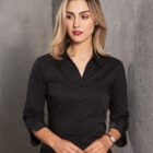 A woman with shoulder-length blonde hair and wearing a black button-up shirt stands against a gray textured wall. She has a thoughtful expression and is holding her hands together. She is wearing a watch on her left wrist.