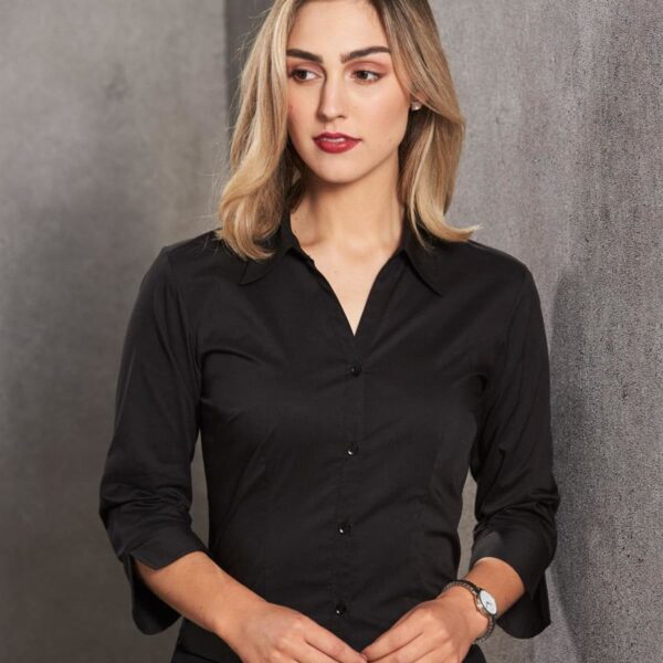A woman with shoulder-length blonde hair stands in front of a textured gray wall. She is wearing a fitted black button-up shirt and a watch on her left wrist. Her hands are clasped in front of her, and she is looking slightly to the side.