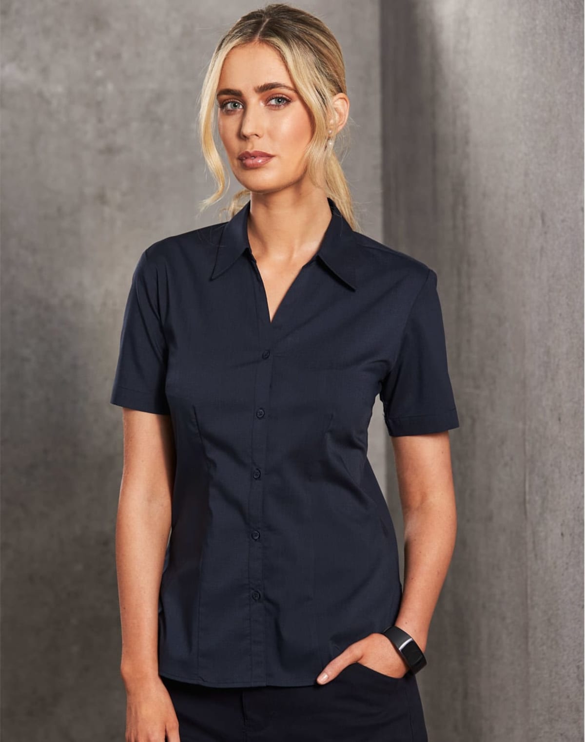 A woman with blonde hair tied back stands against a gray textured wall. She is wearing a dark short-sleeve button-up shirt and dark pants, with a watch on her left wrist. Her expression is neutral, and her right hand rests in her pocket.