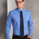 A man wearing glasses is dressed in a blue dress shirt with a black tie and pants. He is standing against a textured gray wall, looking to the side with a neutral expression.