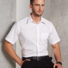 A man with short brown hair and a beard is sitting against a textured wall, wearing a short-sleeve white shirt and black pants. He is looking to the side with a neutral expression.