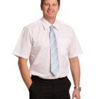 A man in a short-sleeved white shirt and a light blue striped tie stands against a white background. He is wearing black trousers, has short brown hair, and is smiling with one hand in his pocket.