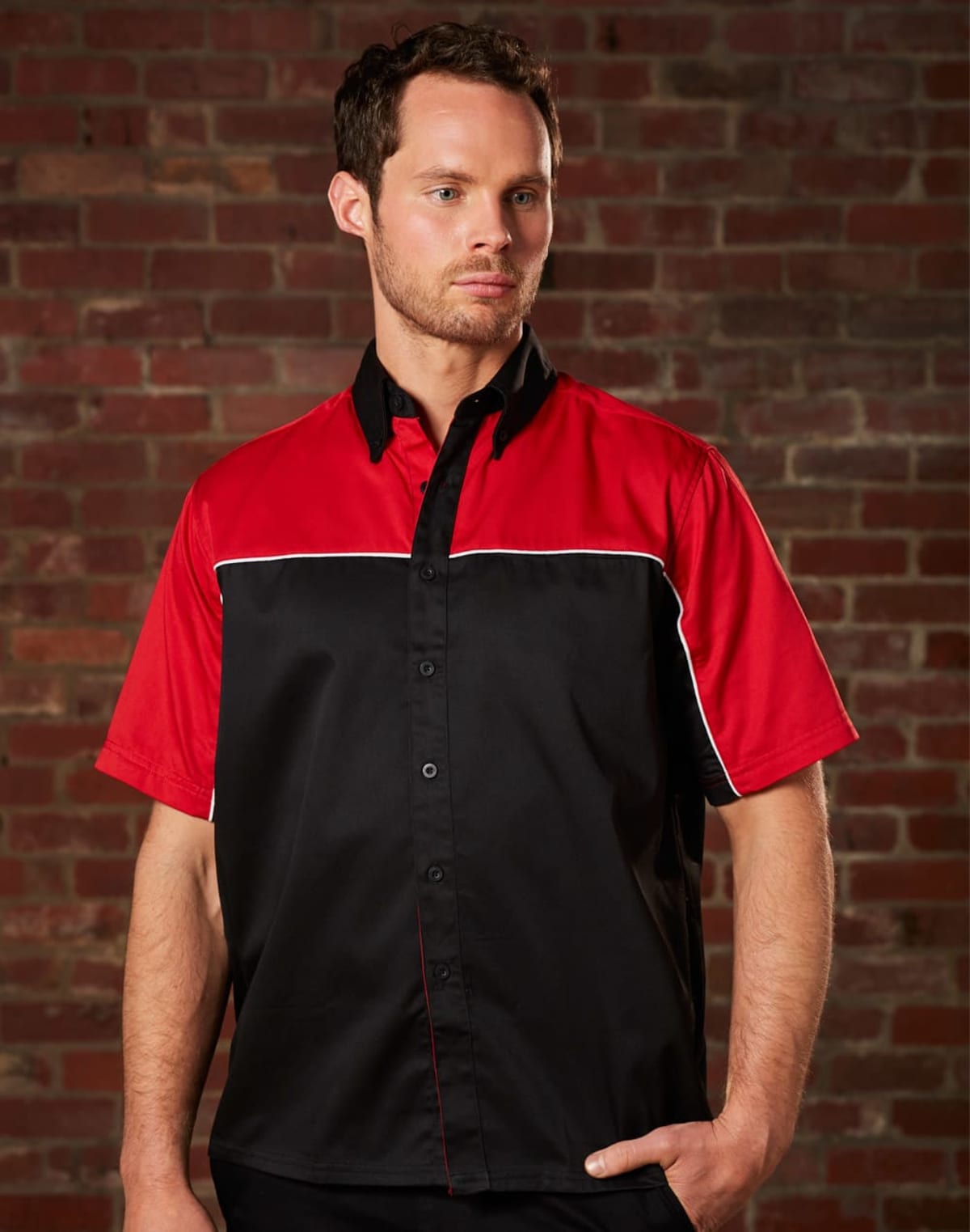 A person wearing a short-sleeved shirt with a red upper section and a black lower section with white piping stands against a textured brick wall. The individual has their hands in their pockets and is looking to the side.