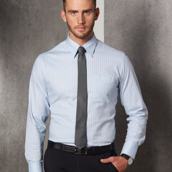 A man wearing a light blue striped dress shirt, dark tie, and black pants stands against a gray background. He has short brown hair and a watch on his left wrist.