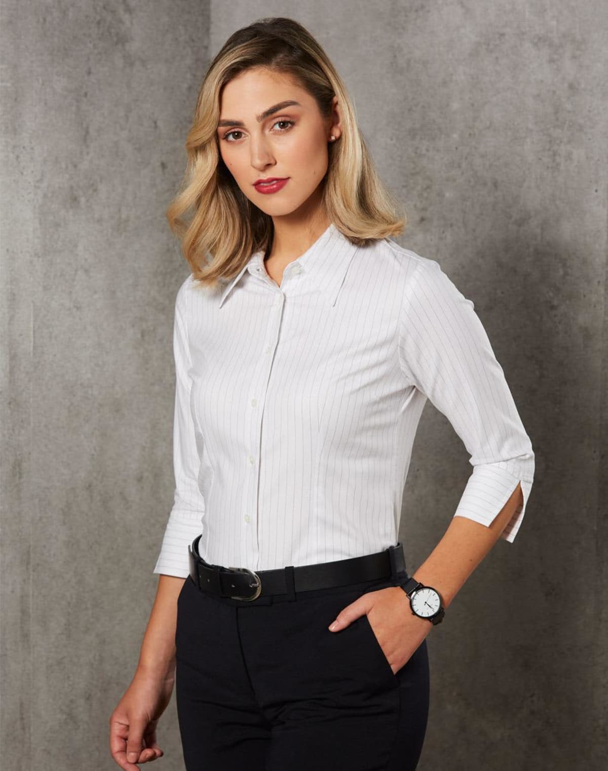 A person with blonde hair wears a white, long-sleeved shirt and black pants, posing confidently with one hand in their pocket against a gray backdrop. They have a watch on their wrist.