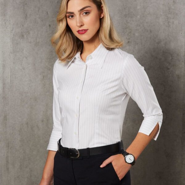 A person with shoulder-length blonde hair is wearing a white, long-sleeved button-up shirt and dark pants. They have one hand in their pocket and are standing against a gray textured background. A watch is visible on their wrist.