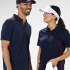 A man and a woman wearing dark blue sports shirts and visors stand side by side, smiling and looking at each other. The woman holds a white towel. The shirts have red accents and both appear relaxed and happy in the casual setting.