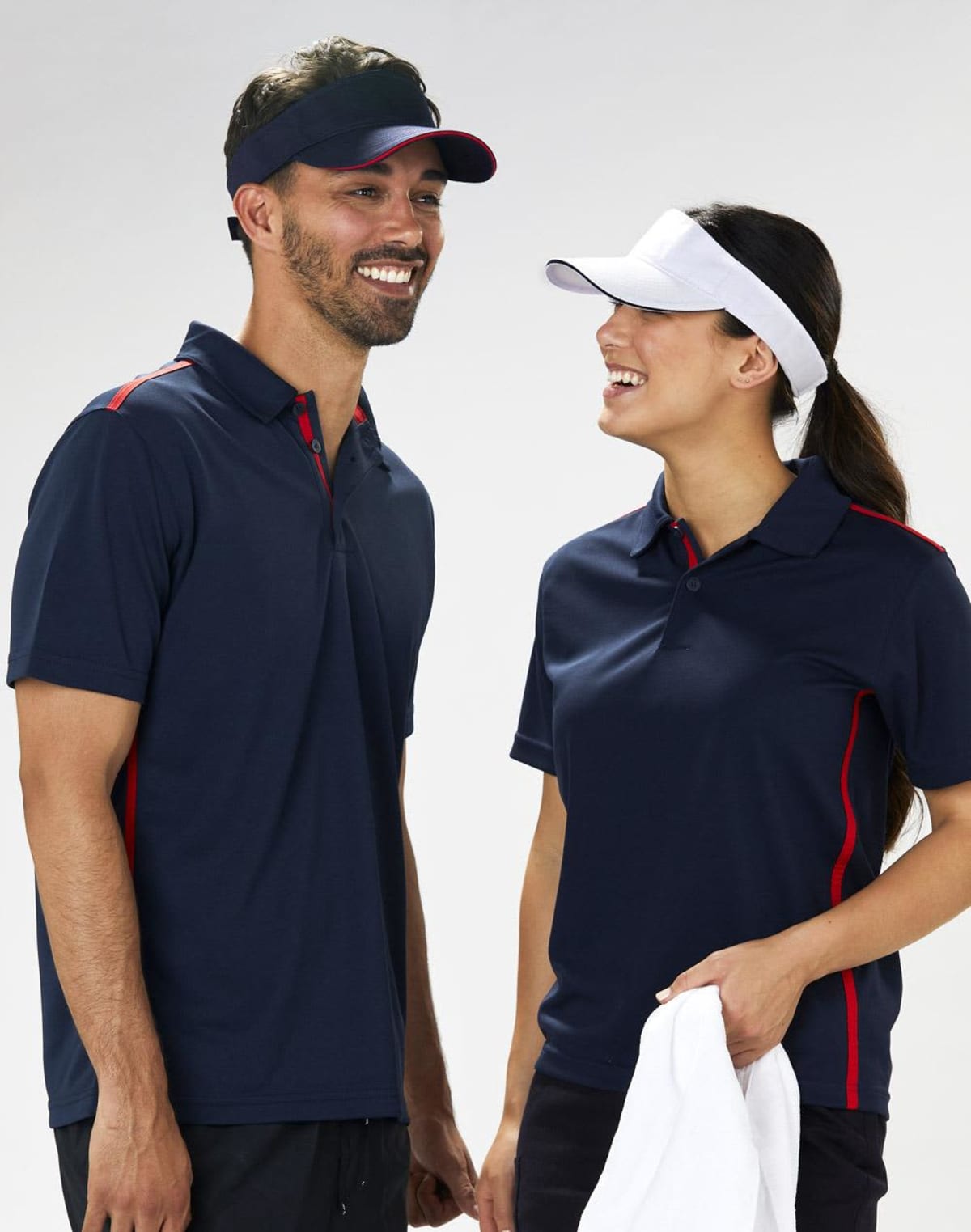 A man and a woman wearing dark blue sports shirts and visors stand side by side, smiling and looking at each other. The woman holds a white towel. The shirts have red accents and both appear relaxed and happy in the casual setting.