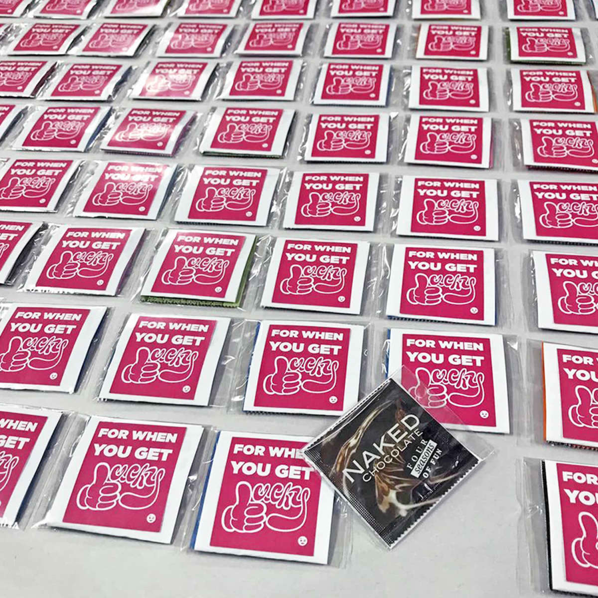 Rows of small packets with a red label reading FOR WHEN YOU GET lucky and a thumbs-up illustration. One packet in the foreground is different, with a black label that says NAKED Chocolate.