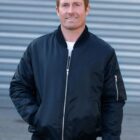 A person with short light brown hair is standing outdoors, wearing a black bomber jacket and a white shirt. They have one hand in their jacket pocket and are smiling at the camera. The background features a metallic, corrugated surface.