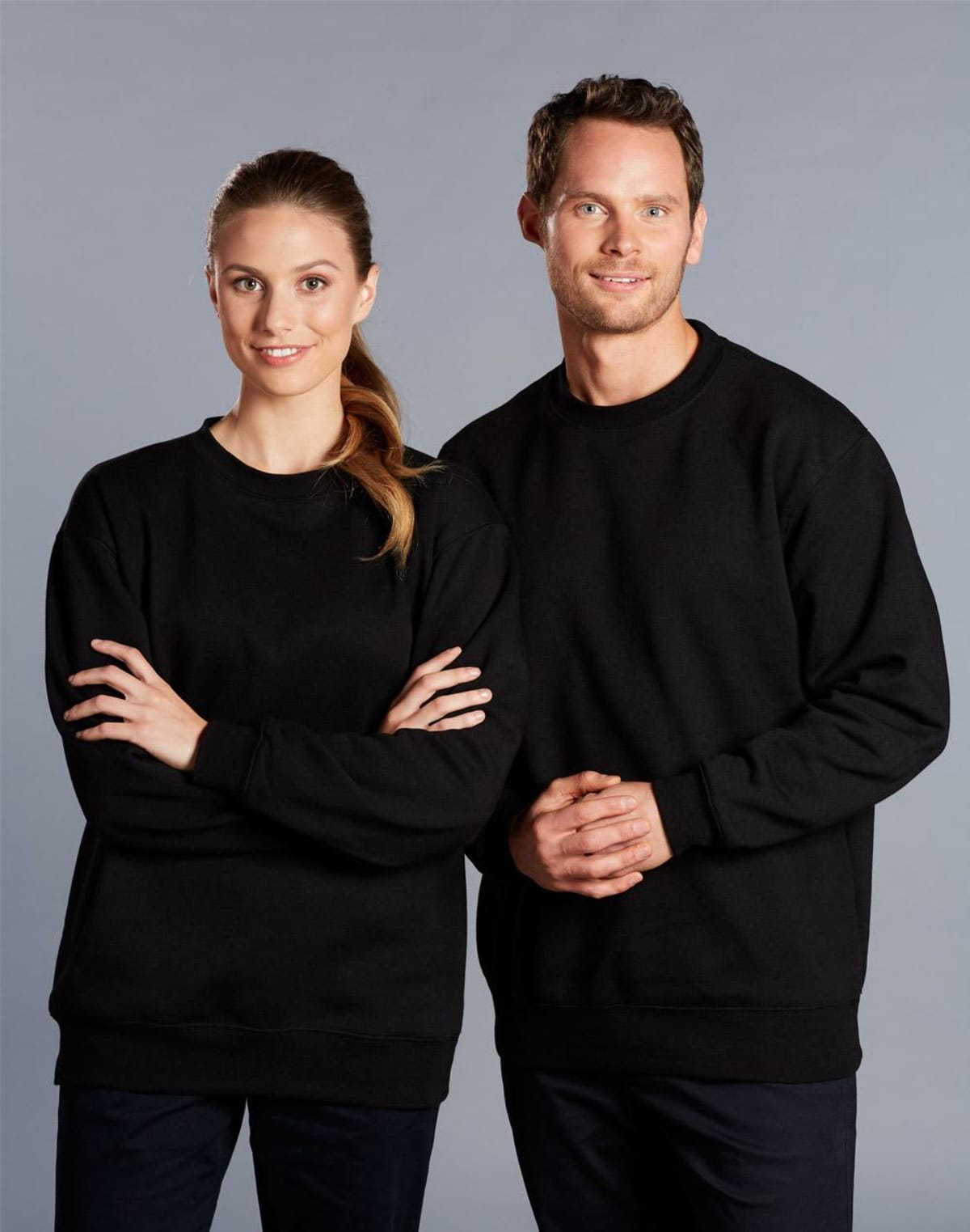 Two people are standing side by side against a gray background. Both are wearing plain black sweatshirts. The person on the left has their arms crossed, and the person on the right has their hands clasped in front.