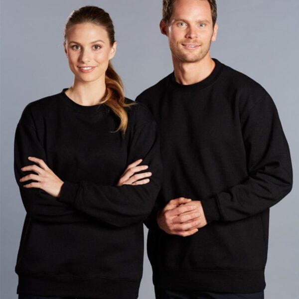 Two people are standing together against a gray background, both wearing black sweatshirts. The person on the left has long hair and their arms crossed, while the person on the right has short hair and their hands clasped in front.