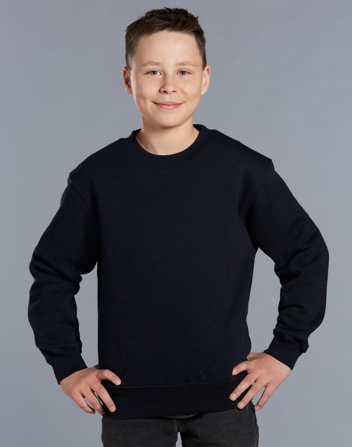 A young boy with short hair smiles confidently, wearing a plain black crewneck sweatshirt and dark pants. He stands against a simple gray background with his hands on his hips.