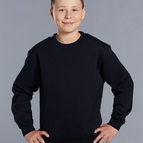 A smiling boy stands against a plain gray background, wearing a black sweatshirt and dark pants, with his hands on his hips.