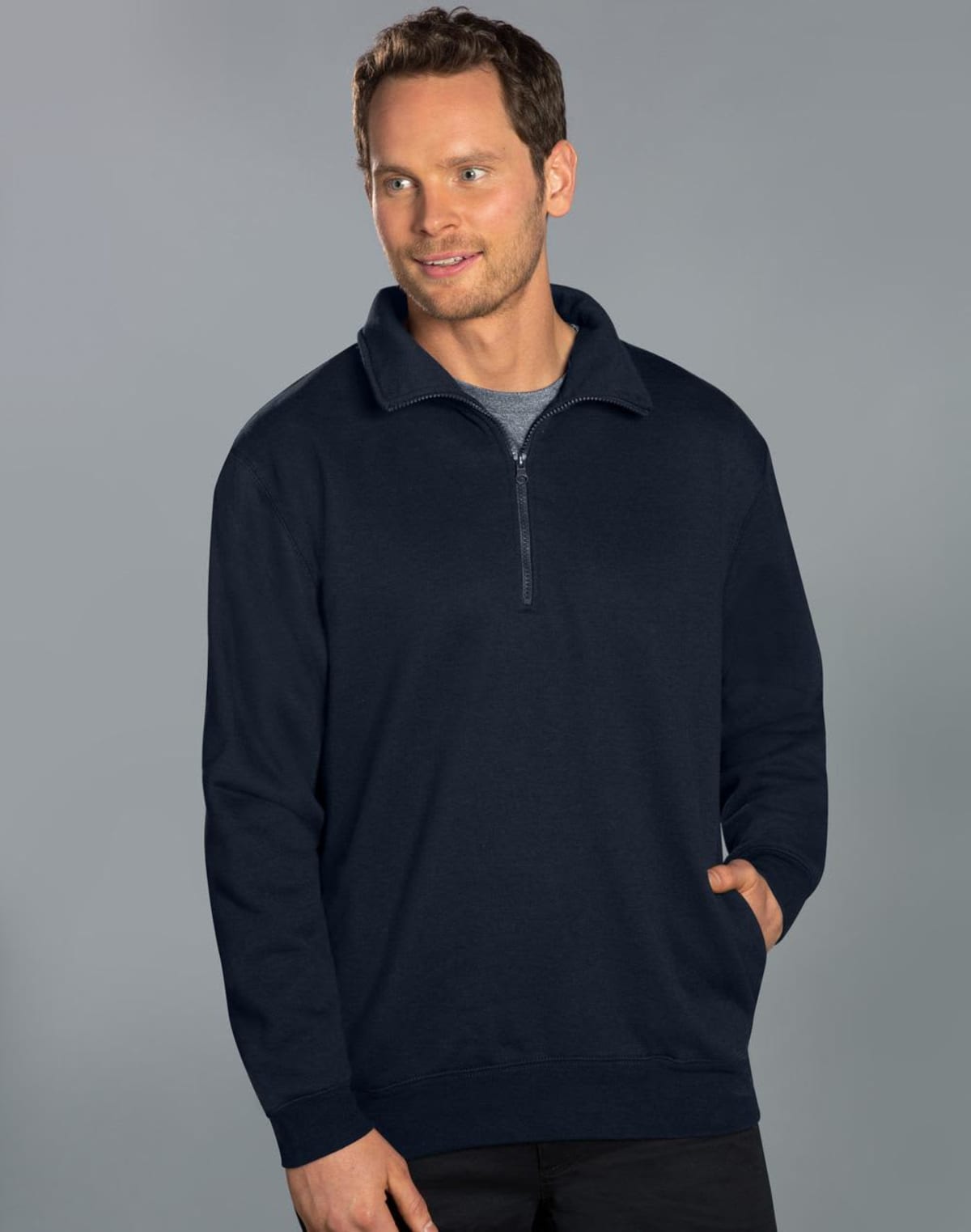 A person stands against a gray background, wearing a navy blue quarter-zip sweater over a gray shirt. They have short hair and are looking to the side with their hands in their pockets.