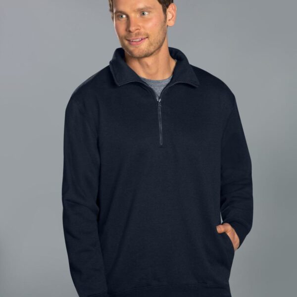 A man wearing a dark blue quarter-zip sweater over a gray t-shirt stands against a plain gray background. He is looking away from the camera with a neutral expression and has one hand in his pocket.