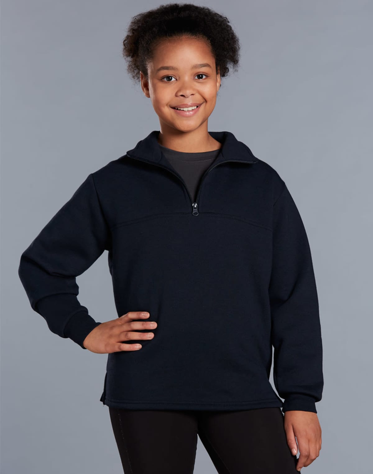A young person with curly hair smiles and stands with one hand on their hip. They are wearing a dark zip-up fleece over a gray shirt and black pants, set against a plain gray background.