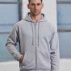 A man in a light gray zip-up hoodie stands with his hand in the pocket against a backdrop of large, blurred windows.