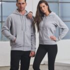 A man and woman stand side by side in a bright room wearing gray zip-up hoodies. The man has his hands in the pockets, while the woman rests her arm on his shoulder. They both wear black pants and are looking forward.