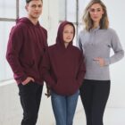 Two adults and a child stand indoors wearing hoodies. The man has a maroon hoodie, the child has a similar maroon hoodie with the hood up, and the woman wears a light gray hoodie. They are standing in front of large windows.