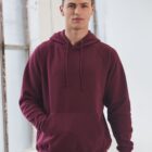 A person wearing a maroon hoodie with a pocket stands indoors near a window. They have short, dark hair and a neutral expression, and they are facing the camera. The background is light and minimalistic.