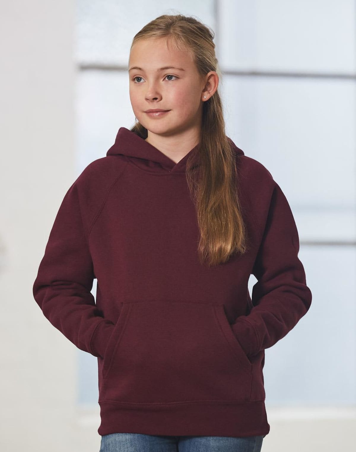 Young girl in a maroon hoodie with hands in the front pocket, standing in front of a softly blurred background featuring large windows. She has long, light brown hair and is looking to the side.