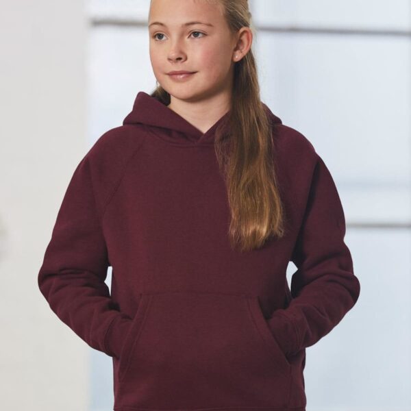 A young girl with long hair wearing a maroon hoodie and jeans stands indoors. Her hands are in the hoodie’s front pocket. She gazes slightly to the side, with a neutral expression. The background is softly blurred, with large windows visible.