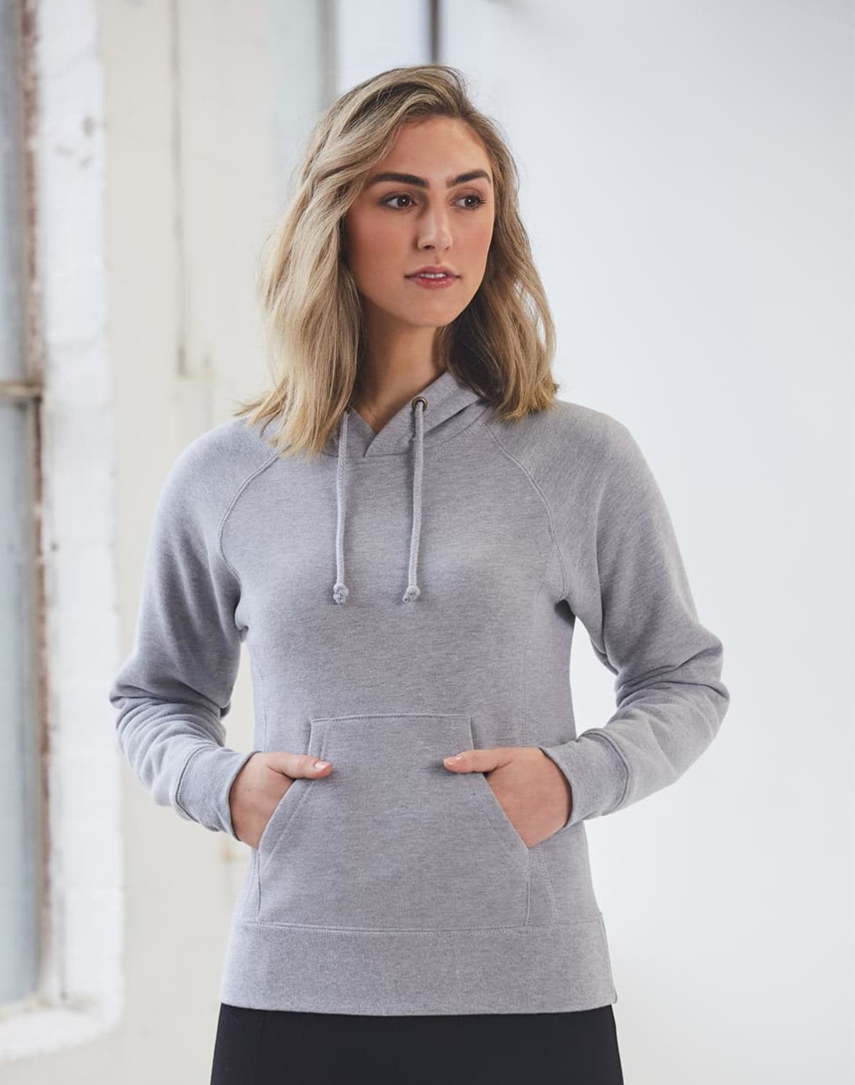 A person wearing a light gray hoodie with both hands in the front pocket stands near a window. They have shoulder-length blond hair and are gazing slightly to the side. The background is softly lit with white walls.