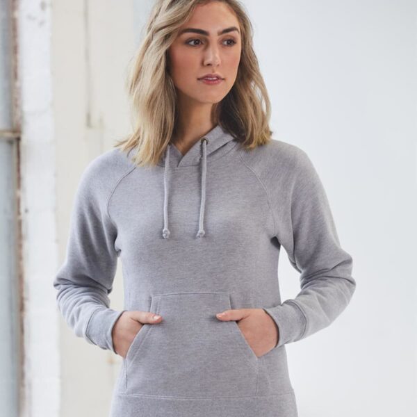 Woman wearing a gray hoodie stands indoors with hands in the front pocket, gazing to the side. Shes in front of a large window with a white frame, creating a bright and airy atmosphere.